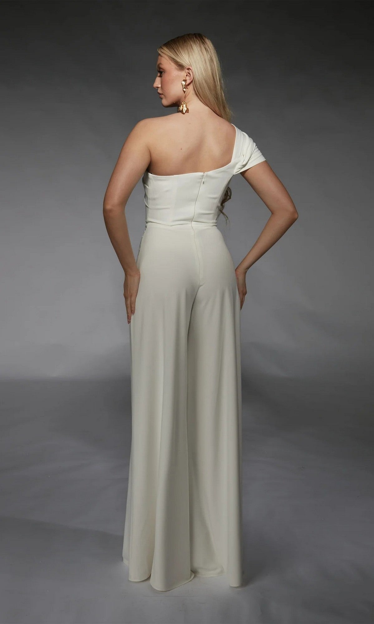 A woman with long blonde hair stands facing away, wearing the Alyce 70066 long formal jumpsuit with wide-leg pants and gold earrings against a neutral backdrop.
